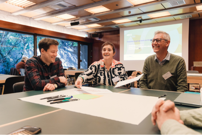 Image showing three participants sitting on a table while doing group work. They are laughing, and there is some paper on the table.